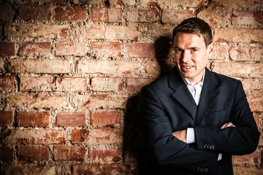Christoph Kleinbeck in a professional suit leaning against a brick wall, representing expert mental performance coaching and leadership.